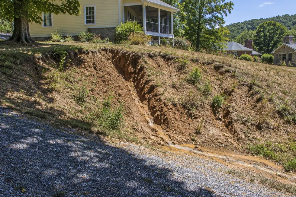 A bare clay bank beside a gravel driveway cut with deep erosion gullies, below a pale yellow clapboard house