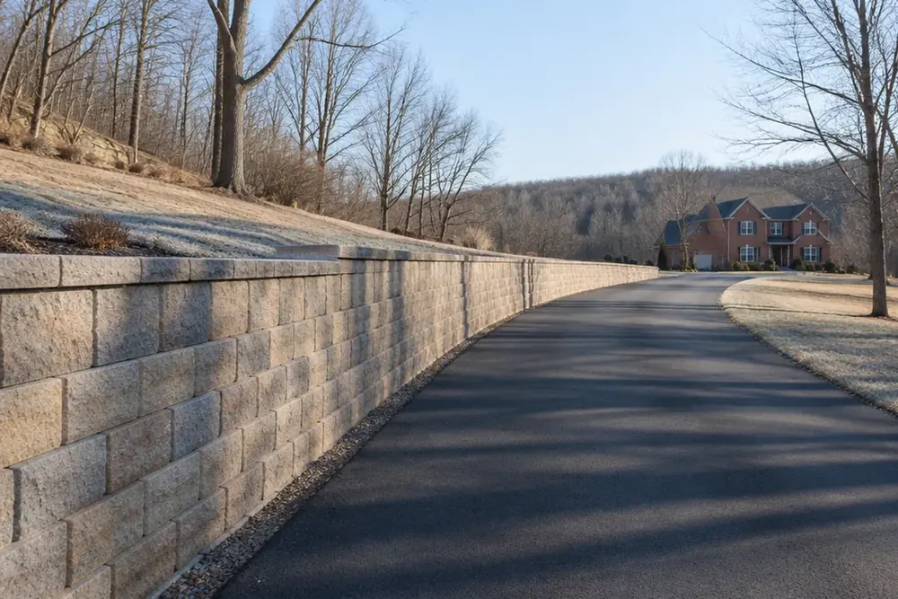A long block retaining wall running away beside a driveway in winter, bare trees behind it